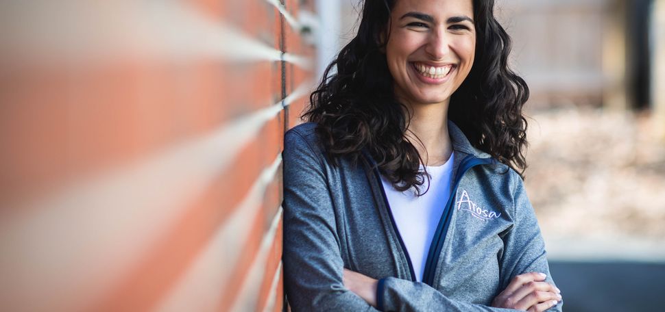 A photo of Gaby Yidi standing against a brick wall with her arms crossed, smiling