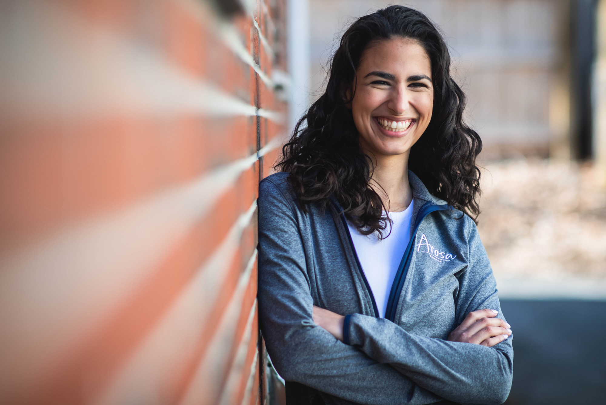 A photo of Gaby Yidi standing against a brick wall with her arms crossed, smiling