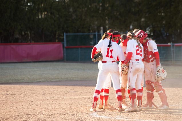 Photo of the women’s softball team in a huddle at the pitcher’s mound; they wear white and red uniforms and look towards the ground as they huddle.