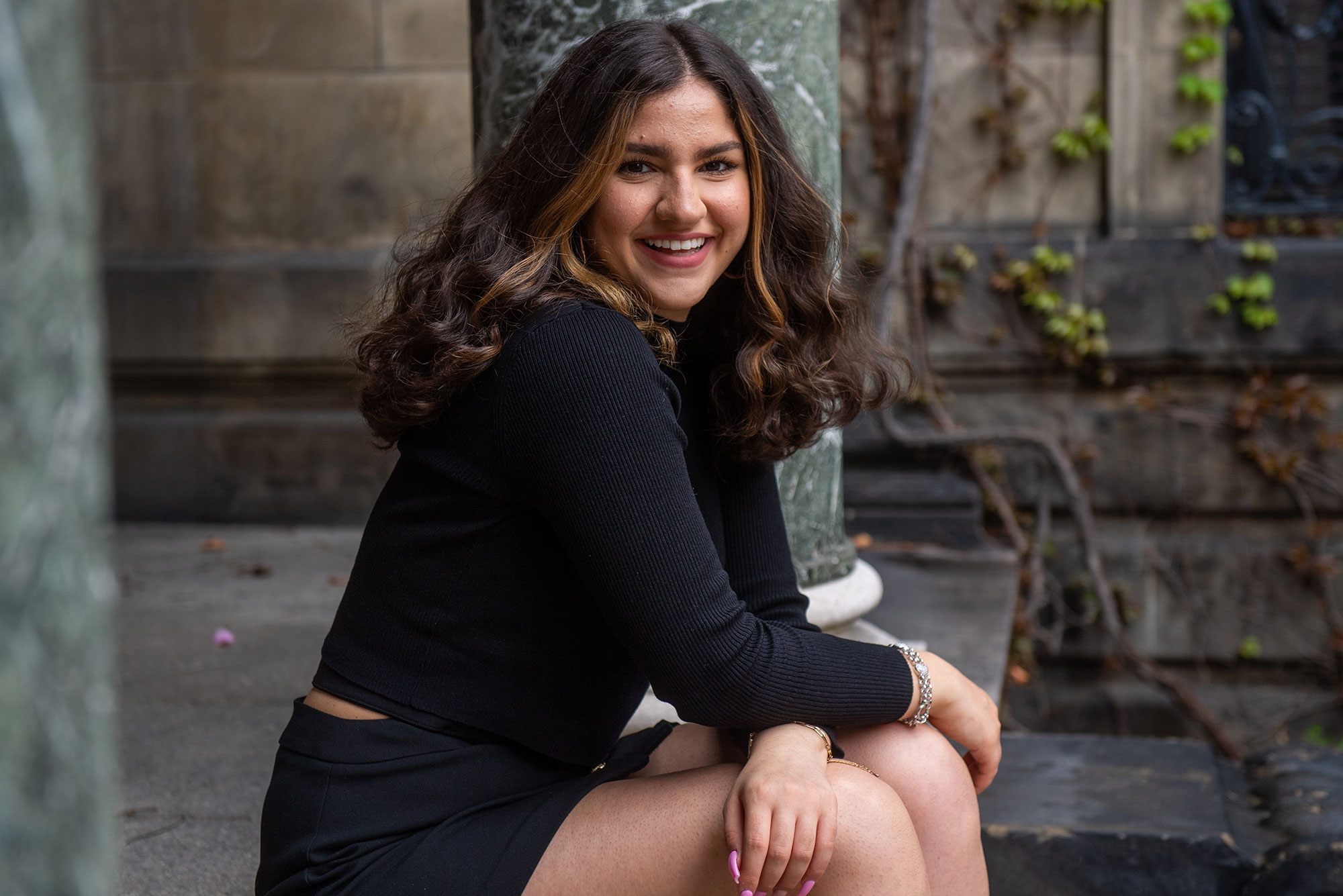 Photo of Tima Dasouki (Pardee’22) sitting on the steps outside of a building with green, granite columns. She wears a black shirt and skirt, has brown shoulder-length hair, and smiles wide.