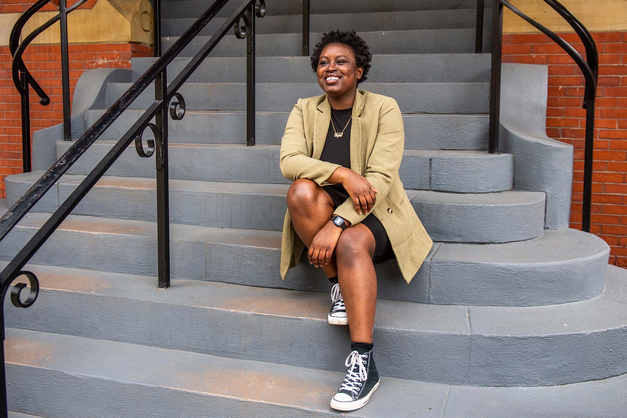 Photo of Cheyenne Bailey (SPH’21) in a tan coat, black dress, and high-top black chucks, sitting on cement steps and smiling.