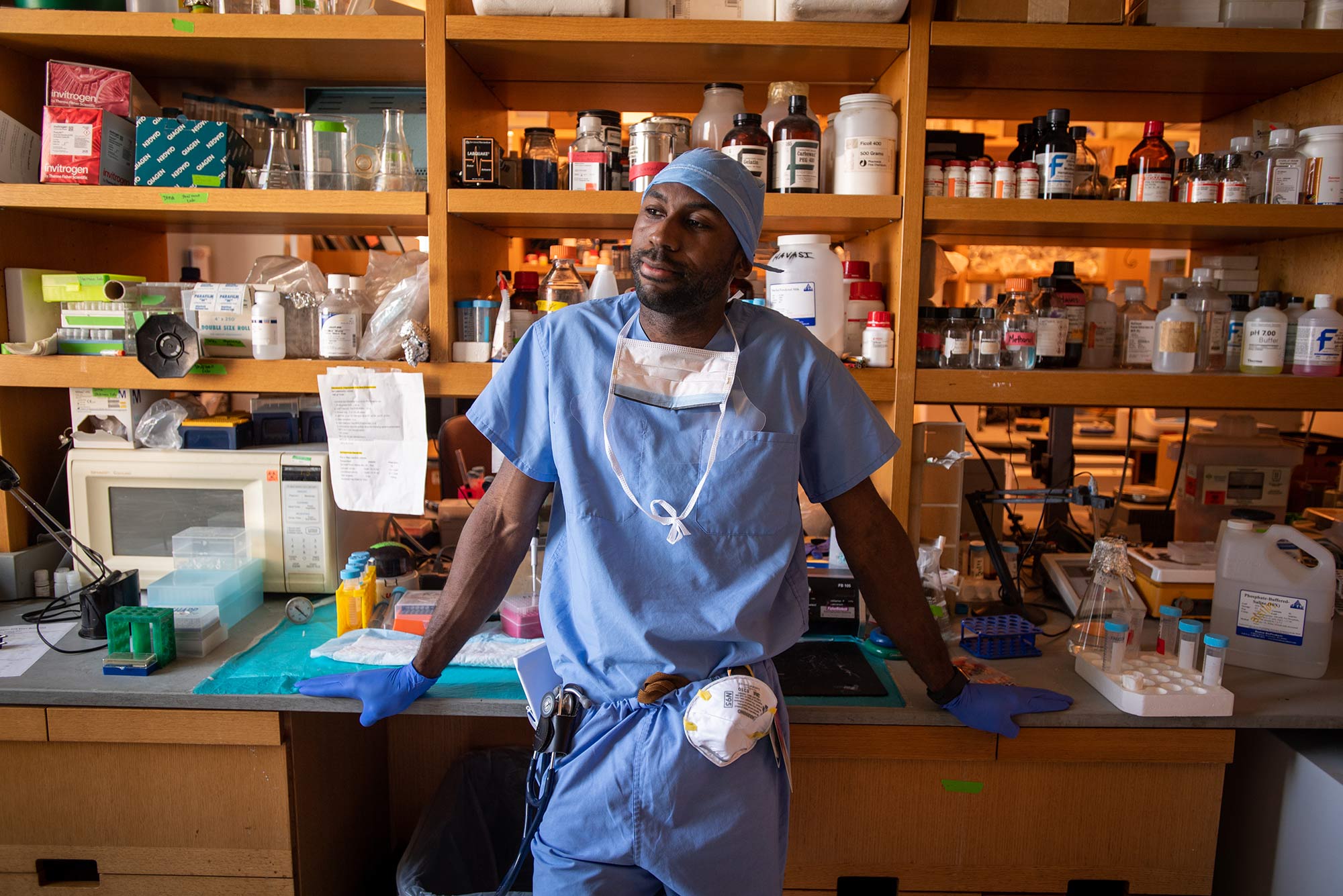 Photo of Chinamere Igwebuike in blue scrubs, blue cap, with his hands resting on a desk behind him, as face masks hang from his neck and from his belt. Behind him, a wooden cabinet is filed with what looks like medical research supplies.