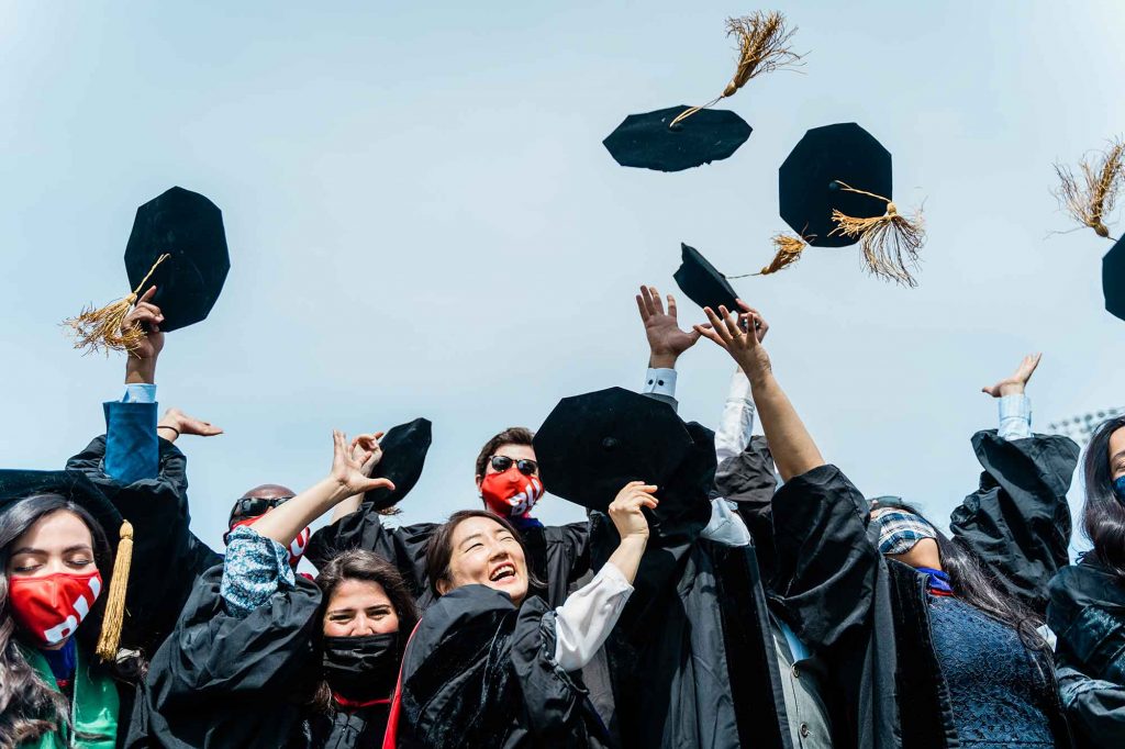 A group of graduating doctoral students wearing black robes and octagonal mortarboards, pose for a photo and throw their caps in the air.