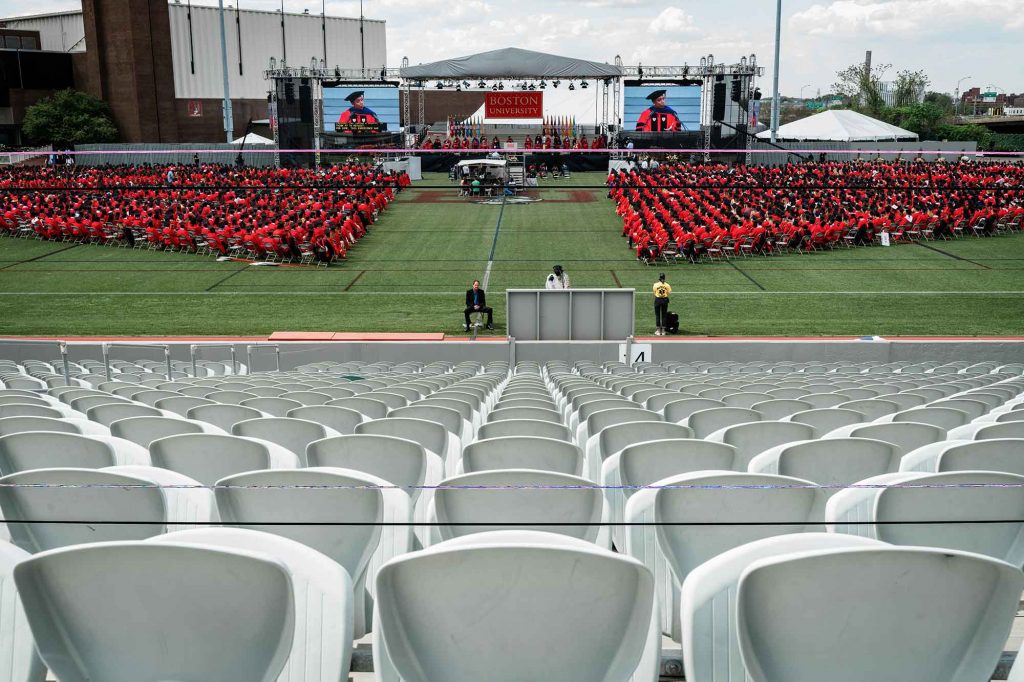 Elevated view of Nickerson Field during the 2021 BU Commencement for undergraduate degrees looking directly center stage from the middle of the bleachers at the back of the field.