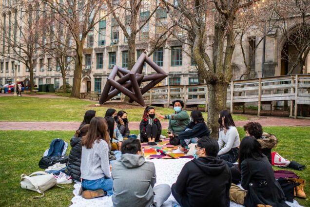 Photo of Swati Rani teaching a writing class on Marsh Plaza on April 6, 2021. The class sits in a circle on blankets on the ground; they all wear face masks. Rani gestures as she speaks as the students listen and look towards her.