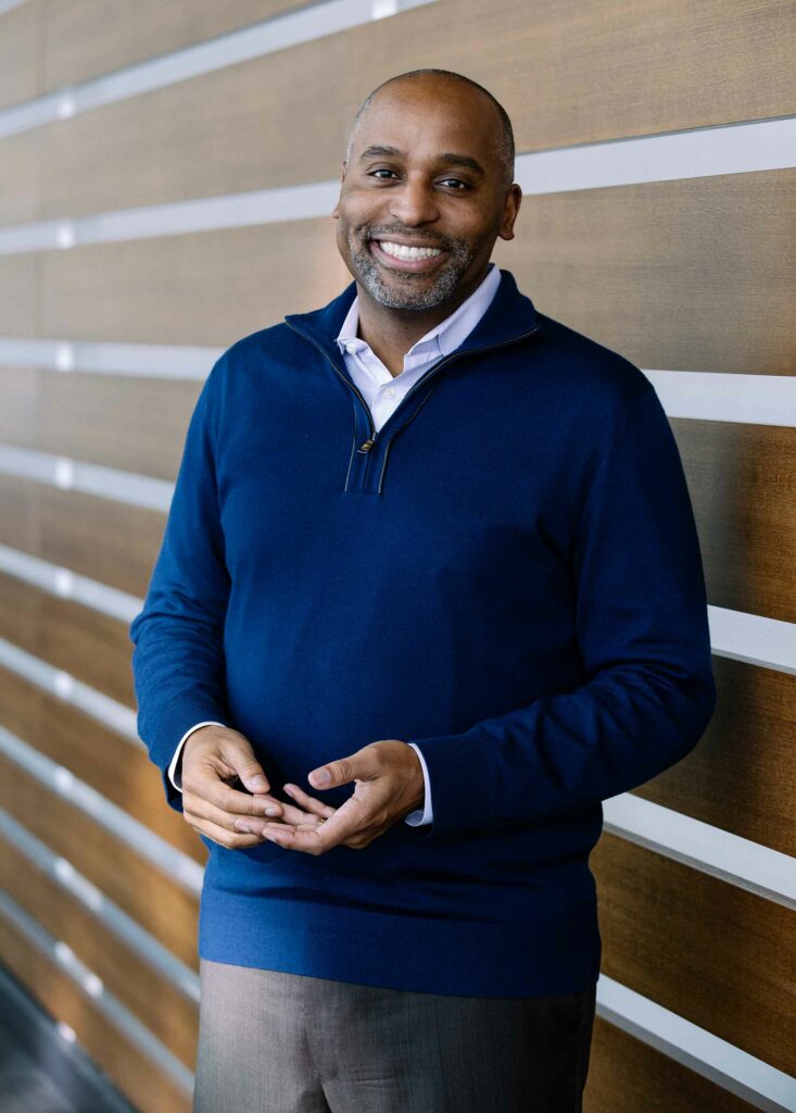 Portrait of CFA Dean Harvey Young in a blue sweater and smiling in front a wall with stripes of wood.