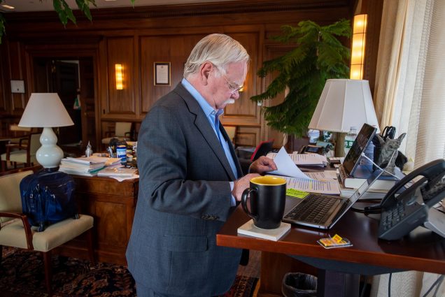 Photo of President Robert A. Brown looking at a papers on his desk, in front of his laptop, as he prepares for the Wednesday morning town hall via Zoom in his office. He wears a blue suit; his wood-paneled office is seen in the background.