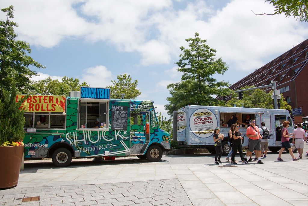 Photo of two food trucks parked along the Greenway, on the left, a green and blue truck with stripes called ‘Shuck’ is parked, while on the right, a light blue truck called ‘Cookie Monstah’ is parked. People stand and walk by in front of the latter.