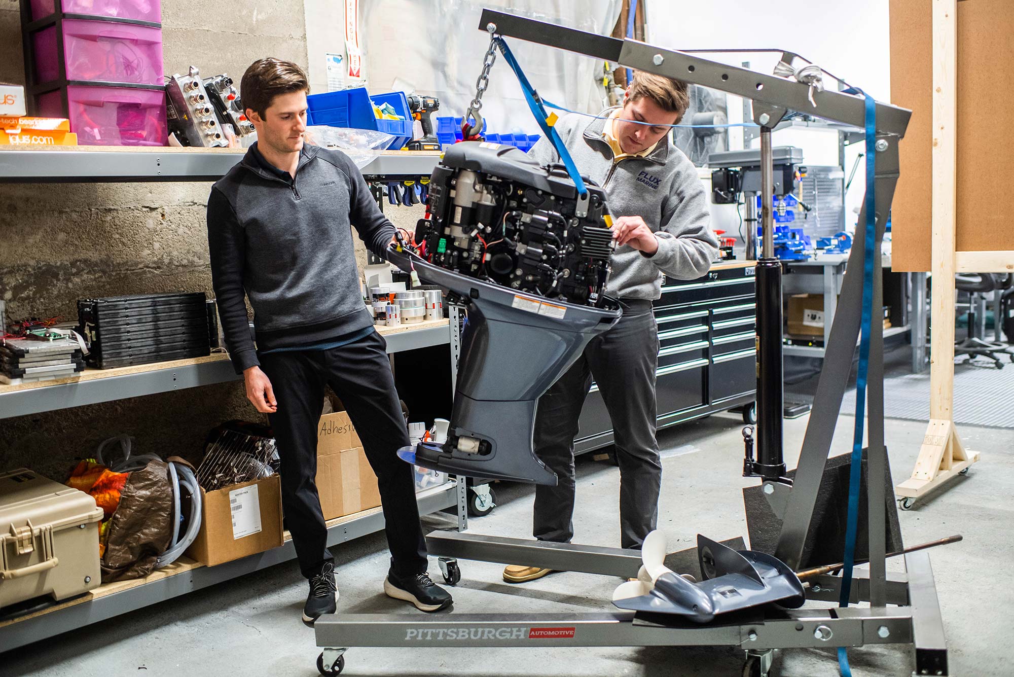 Photo of Daylin Frantin, left, in a gray long-sleeve and black pants, with one hand on a boat engine, while Sam Gallipeau, Flux’s in-house designer, in a gray quarter zip, inspects the other side of the engine. Behind them are work benches and industrial shelves. A small crane lifts the engine