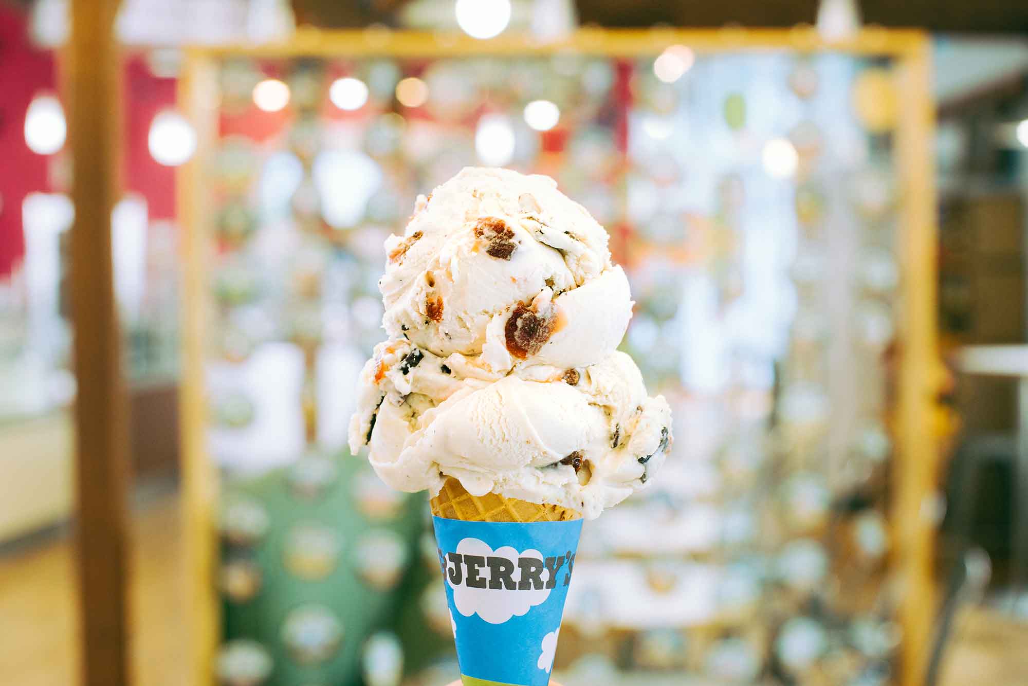 A Cherry Garcia ice cream cone is held up against the backdrop of a Ben and Jerry's brick and mortar ice cream shop.
