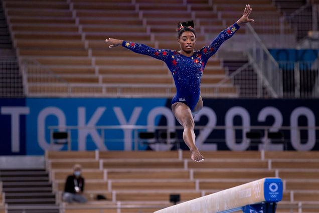 Photo of Simone Biles, a Team USA gymnast, with her arms raised at her sides, and her legs kicking out, as she jumps above the balance beam before empty stands at the Ariake Gymnastics Centre during the 2020 Tokyo Summer Olympics, Sunday, July 25, 2021.