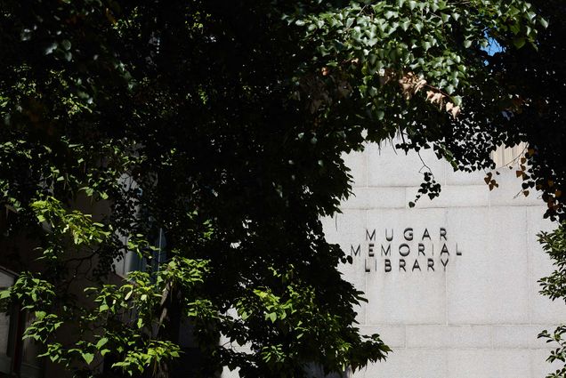 Photo of the entrance to the library as seen through a break in the trees. A sign that reads “Mugar Memorial Library” hangs on a gray, granite wall.