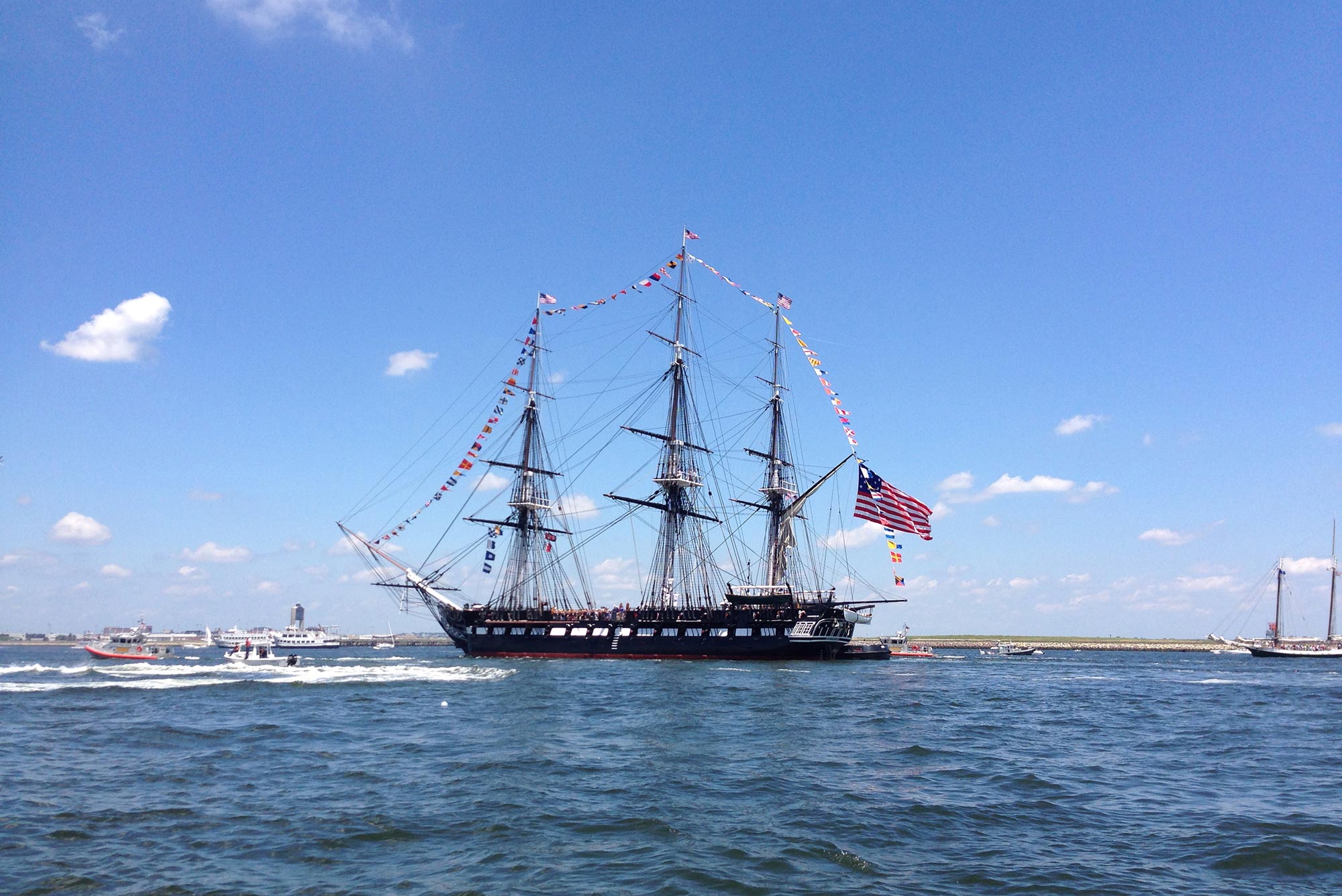 Photo of the USS Constitution (also known as “Old Ironsides”) in the harbor on a summer day with a few small clouds in the sky. The ship is decorated for the fourth.