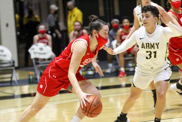 Photo of Riley Childs (COM’22) dribbling a basketball game during a game against the team “ARMY” in white jerseys, a defender stands with their hands up behind her to her left. Childs wears a red face mask.