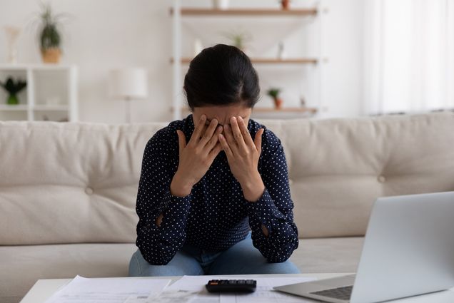 Photo of a young woman with her face in her hands as she sits on a couch in front of a laptop.