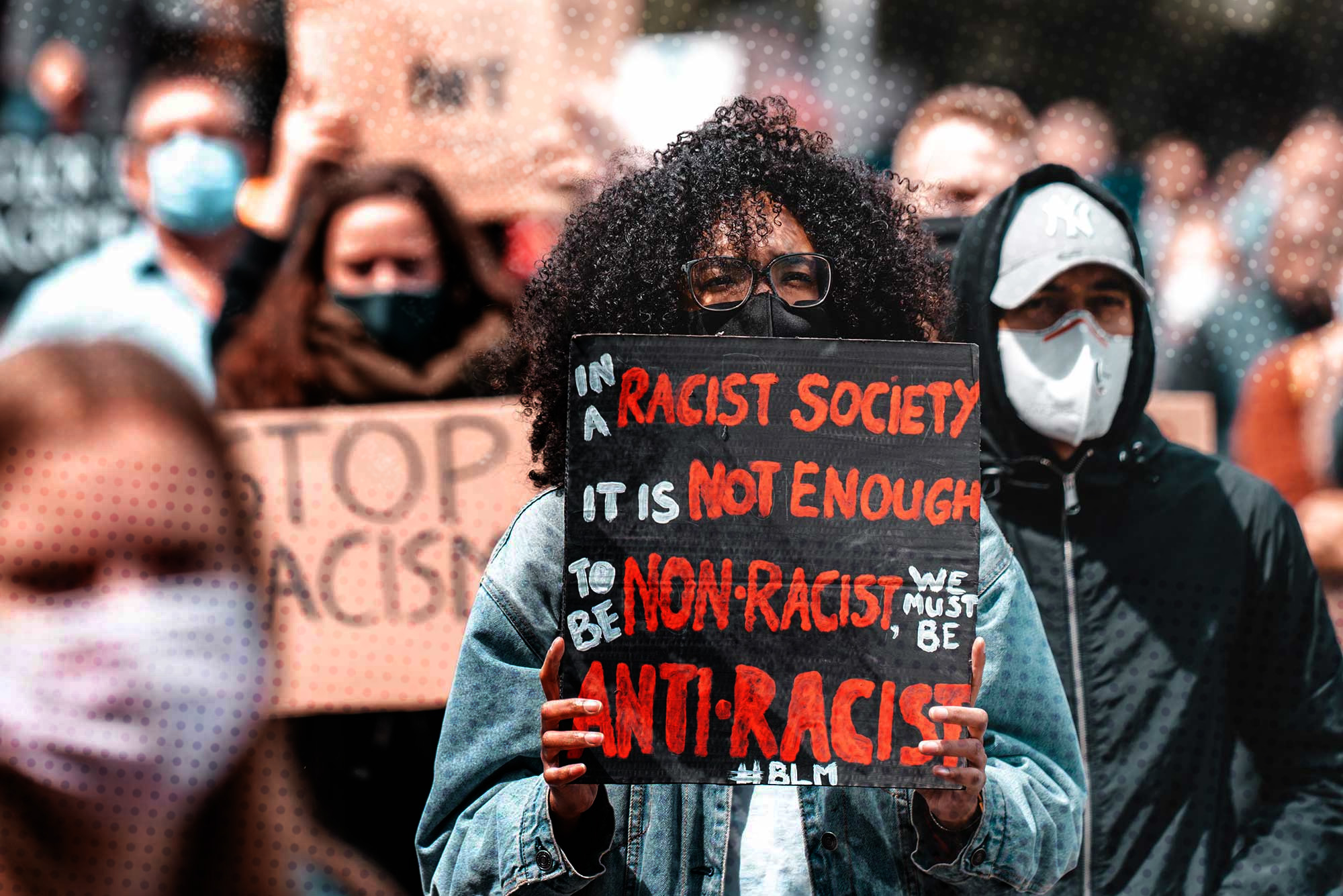 Photo of an African American woman holding a sign that says "In a racist society, it's not enough to be non-racist. We must be anti-racist. #blm" at a protest