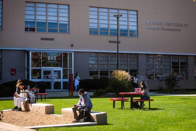 Students wearing protective face coverings sit at tables and benches on the lawn outside the Boston University College of Communication