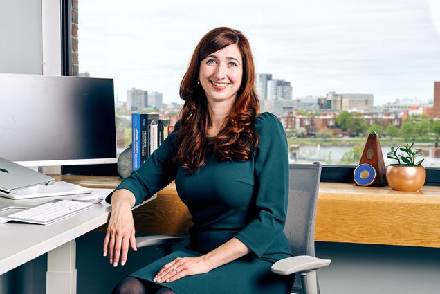 Photo of neuroscientist and speech pathologist, Jenny Zuk, in a hunter green dress, sitting at her desk with her arm resting on the desk. Behind her, the Boston skyline is seen.