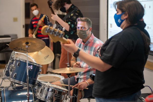Photo of Paul Halpainy (CFA’19) in rock band performance and pedagogy at CFA August 2. He is seated at the drum, and a person with a black t-shirt plays the bass guitar in the right foreground.