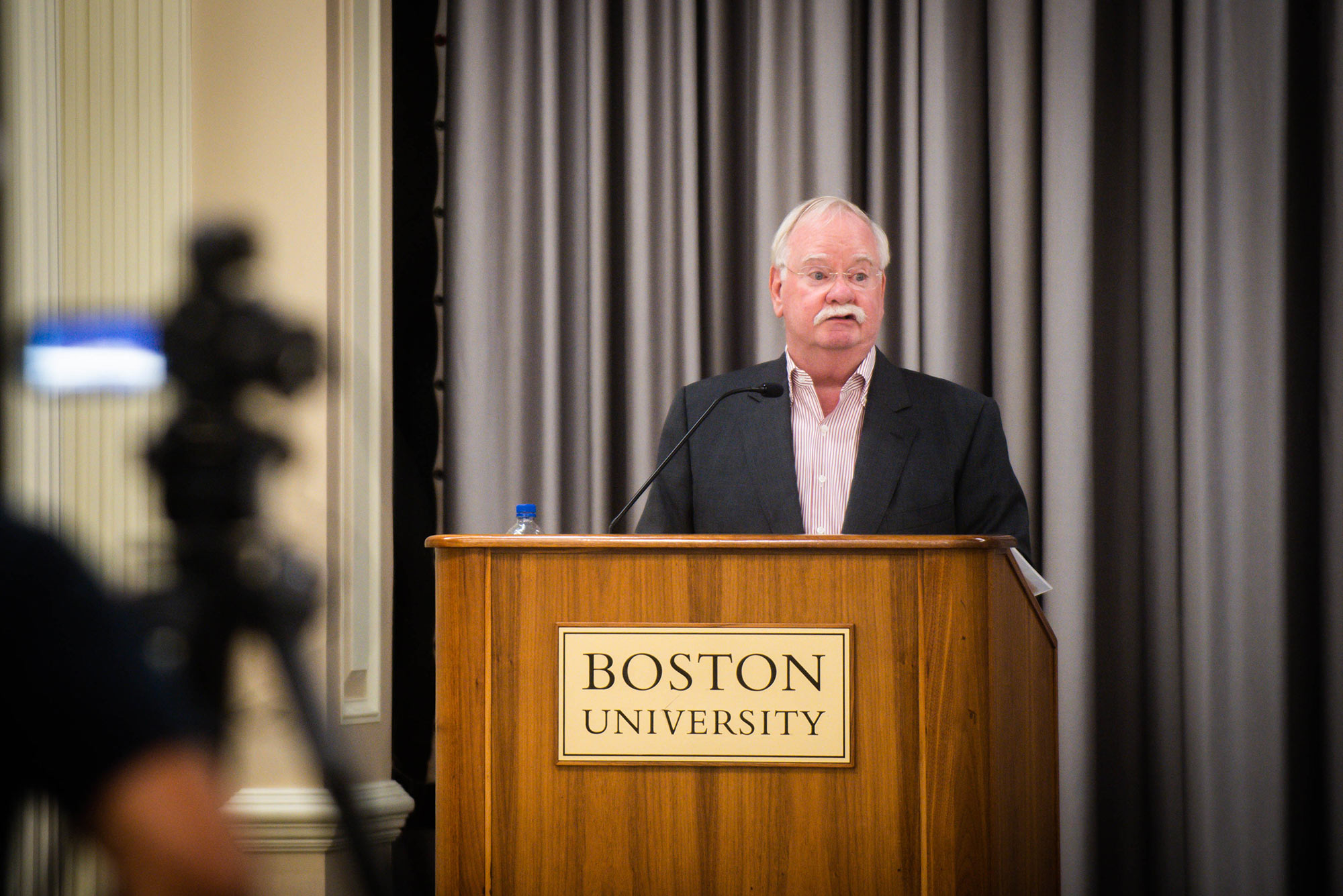 Photo of Dr. Robert Brown, in a suit, standing at a Boston University podium, discussing the fall semester and the beginning of remote work during a town hall meeting on August 19, 2021. A person filming the event is seen in silhouette in the left foreground.