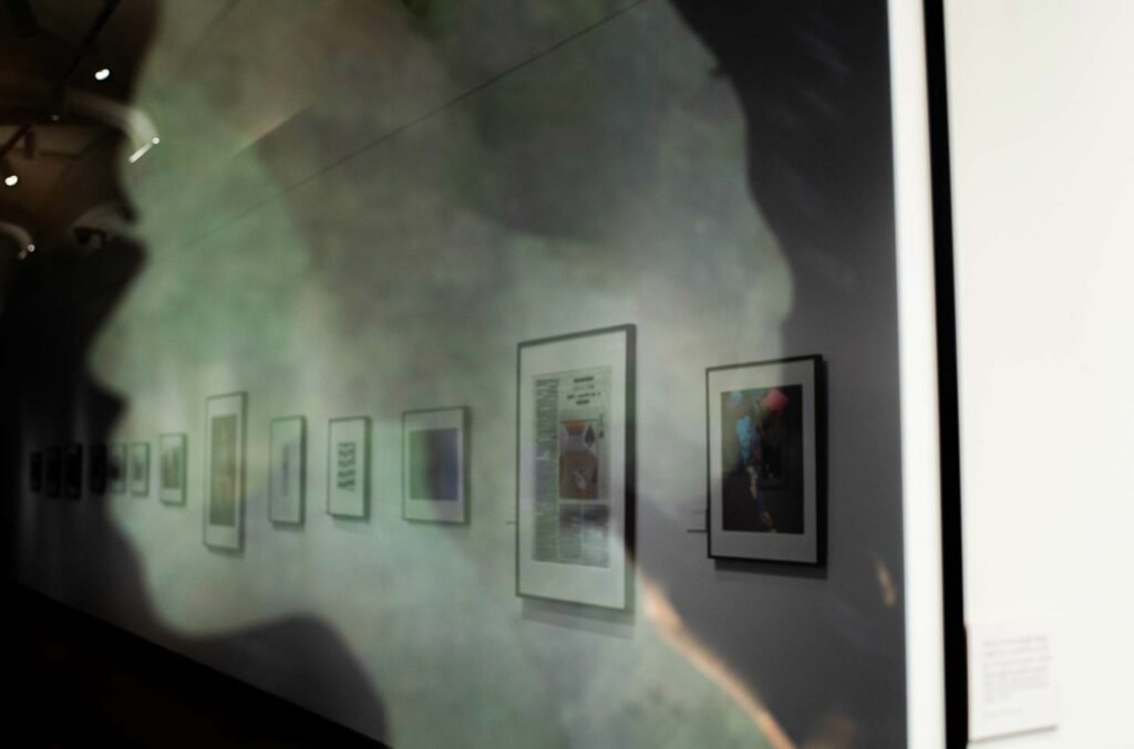 Photographs in the A Yellow Rose Project exhibit are seen hanging on the wall of Stone Gallery behind the silhouette of a woman's profile in the reflection of frame glass.