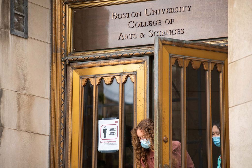 Students exit the Boston University College of Arts and Sciences building wearing protective face coverings. A sign on the door to the building notifies people that face masks are required inside.