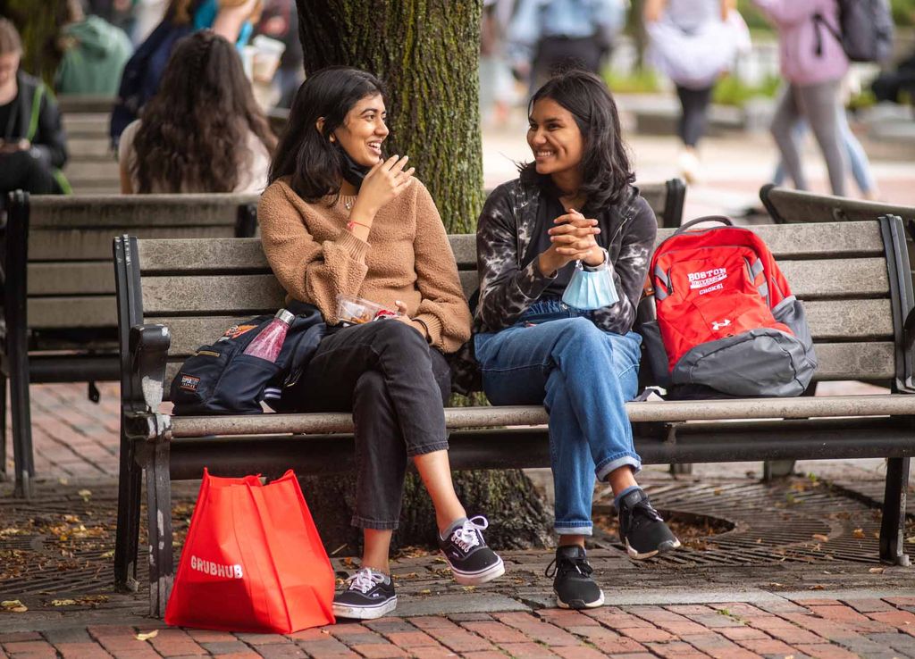 Two students with red Boston University backpacks sit on a bench talking.