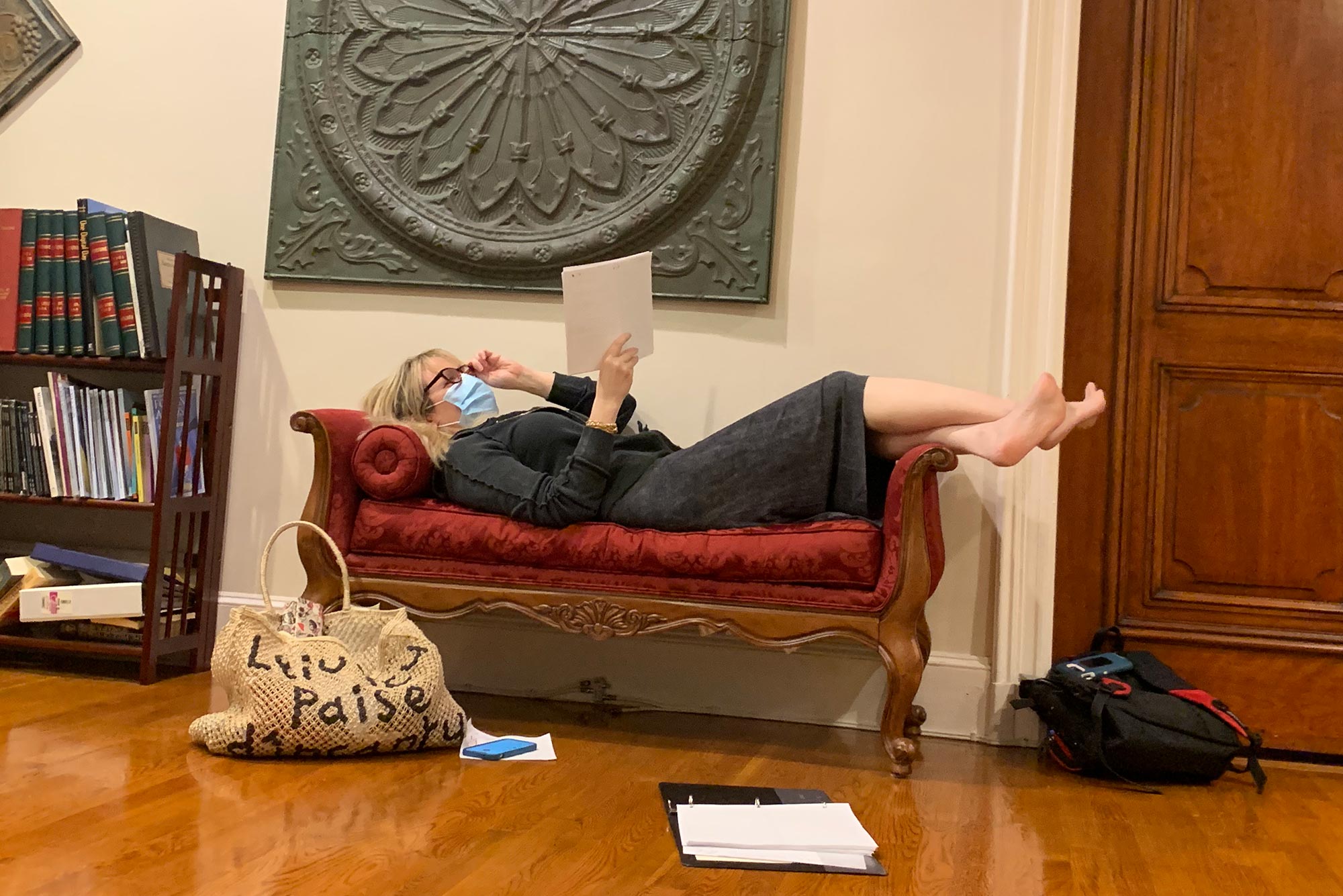 Photo of Wendy Lippe (GRS’93,’96), here rehearsing for the Psych Drama Company’s audio productions. She wears a knee-length gray dress and a blue face mask and reclines on a red therapy-looking couch with a script in her hand.