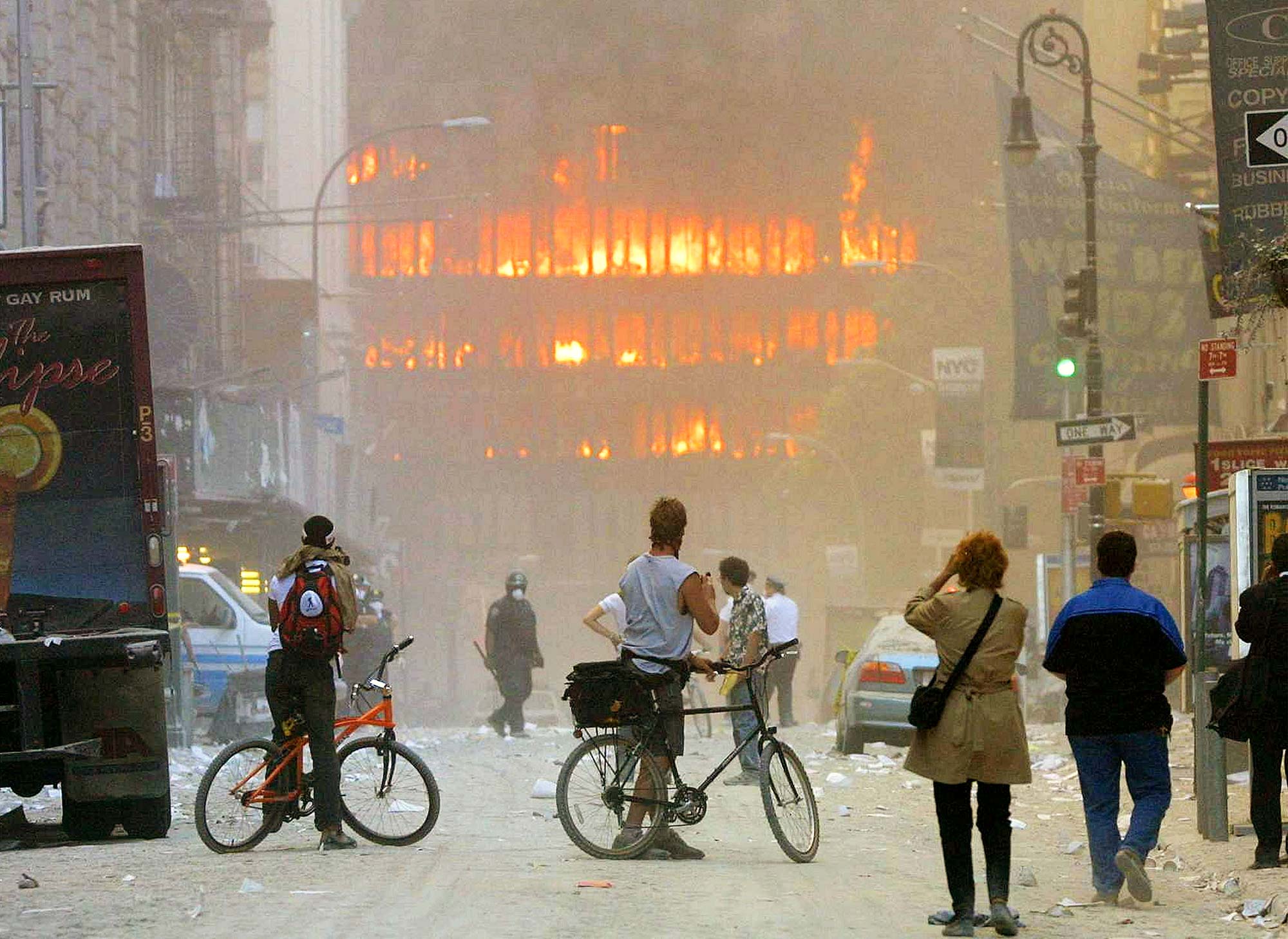 Photo of two people on bikes and other bystanders looking towards the twin towers as they burn. The air is filled with ash and the photo is very hazy.