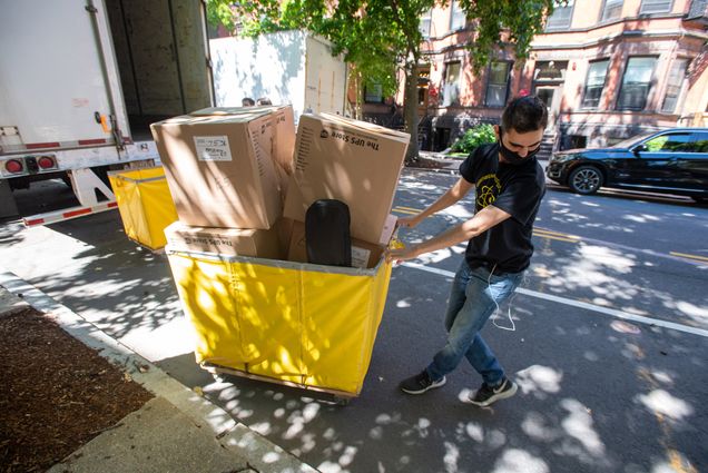 Photo of August Menchaca (CAS’23) in a face mask, black t-shirt and jeans, collects his things from a UPS truck and wheels them away in a yellow cart while moving in on Bay State Rd on August 26.