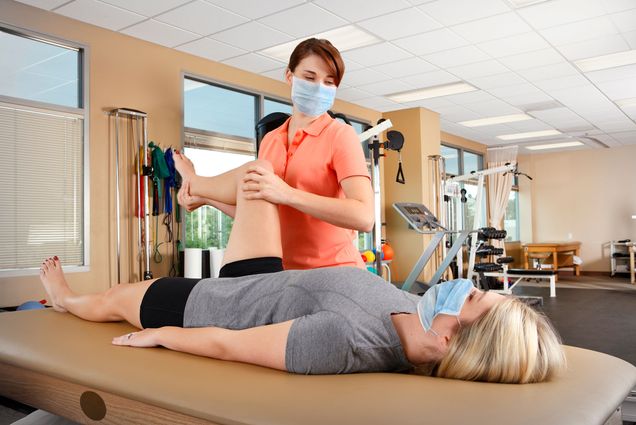 photo of a physical therapist, wearing a protective face mask, evaluates the range of motion of a young female patient's hip as she lies on a treatment table. The scene is inside a physical therapy clinic with several pieces of exercise equipment in the back