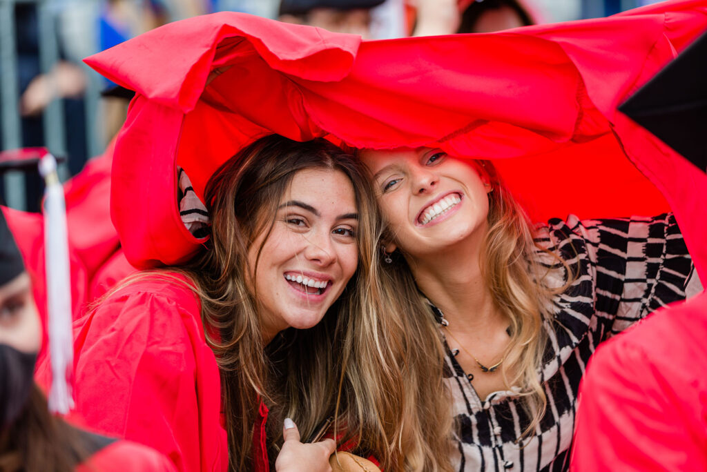Photo of two young white women at 2020 Commencement. They smile as they hide from the rain under one woman's red gown.