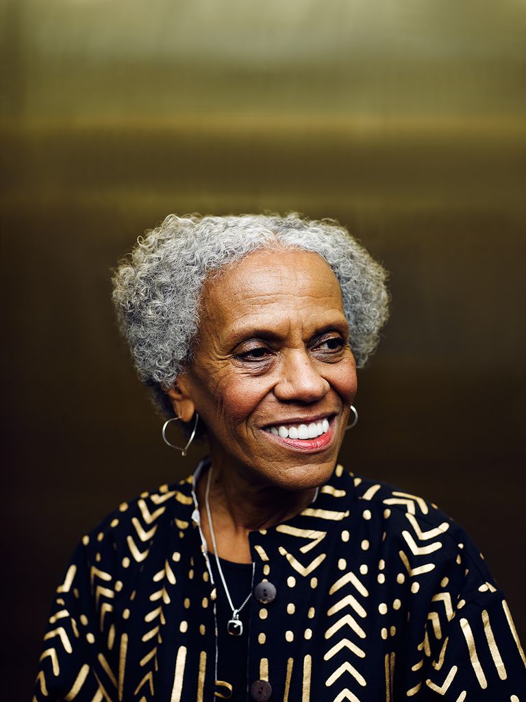 smiling headshot of Andrea Taylor. She looks to the right in front of a golden metal background. She wears a black and gold shirt.