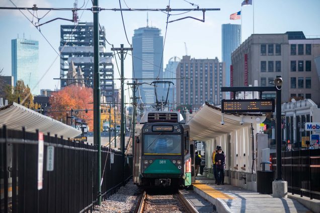 photo of an eastbound Green Line trolley pulling into the MBTA’s new Amory Street station. Two people wait under the new overhang of the station as the green trolley pulls up on the tracks.