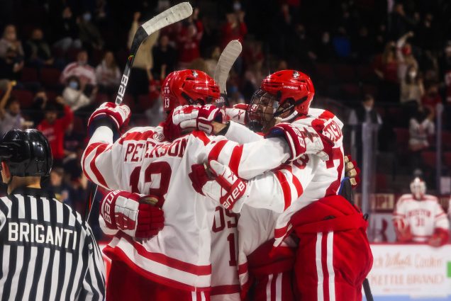 Photo of BU men’s ice hockey team celebrating a goal in its 2-2 tie against UMass Amherst at Agganis Arena on November 12. The men wear white and red jersey and hold their sticks up straight as they hug.
