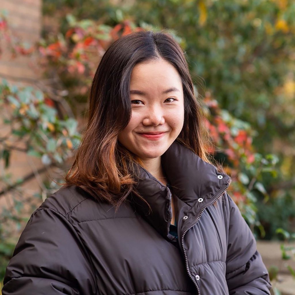 photo of a smiling Kristi Li standing in front of foliage on BU's campus. She wears a black puffer jacket and stand with hands in pockets.