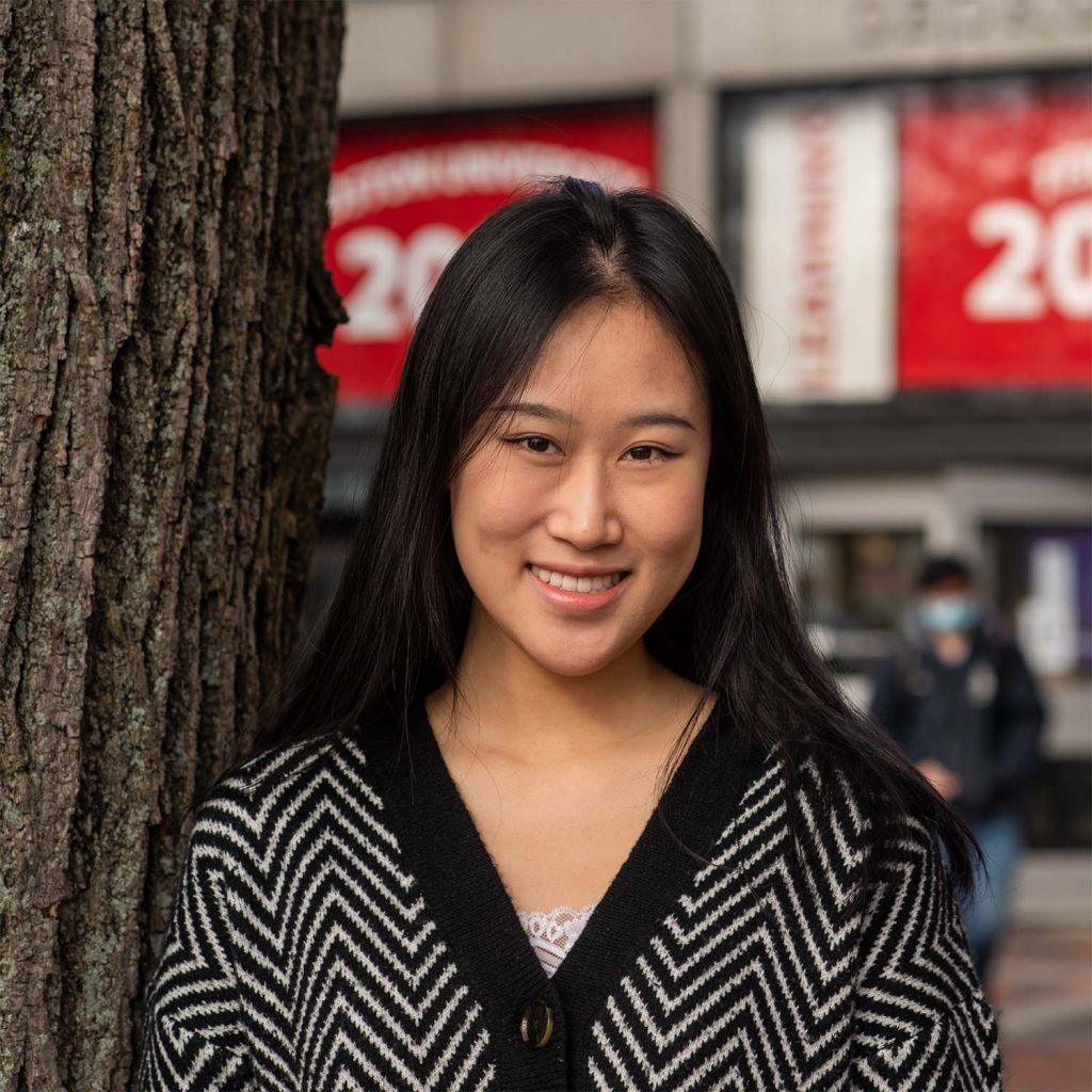 photo of a smiling Yuli Wen leaning against a tree in the center of BU's campus. She wears a black and white striped, button-up sweater.