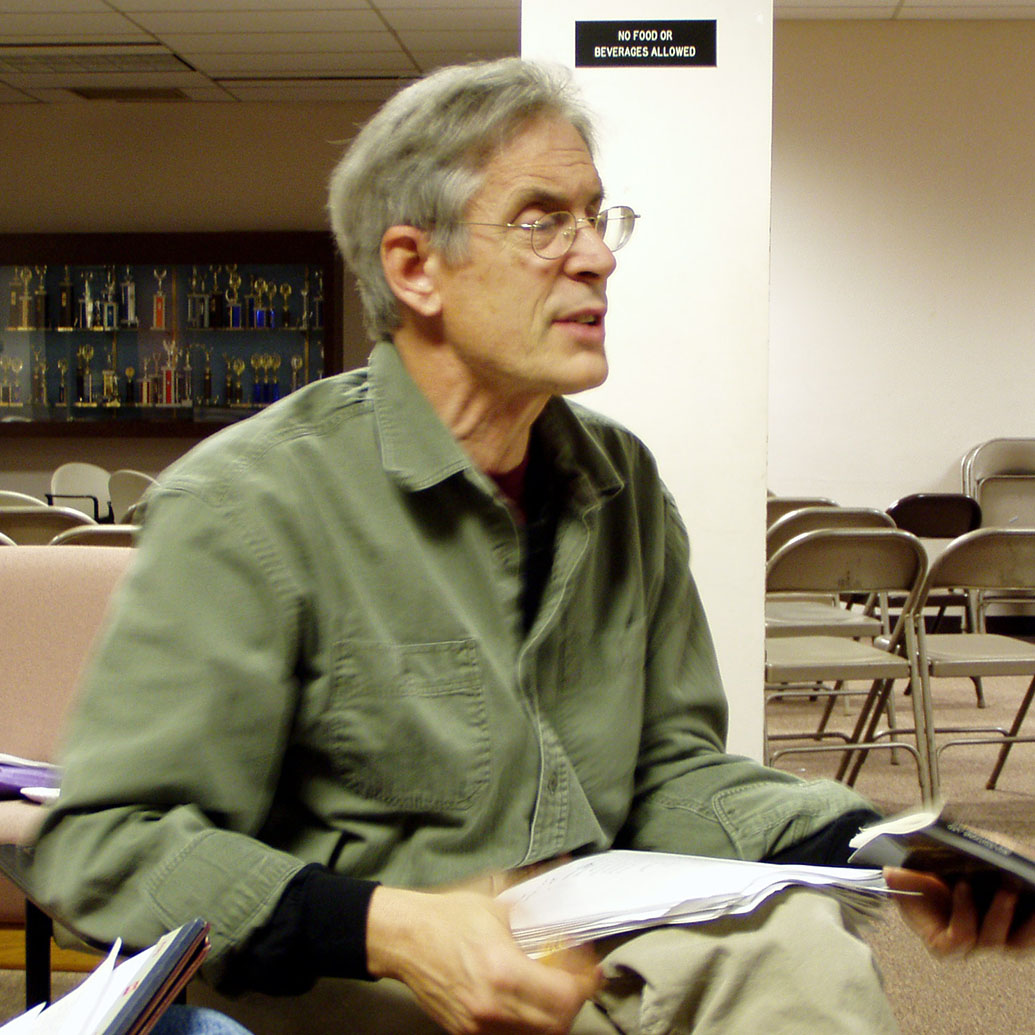 Candid photo of  award-winning poet Christopher Bursk teaching a class. He wears an olive-green shirt and holds a book in hand as he speaks to his class