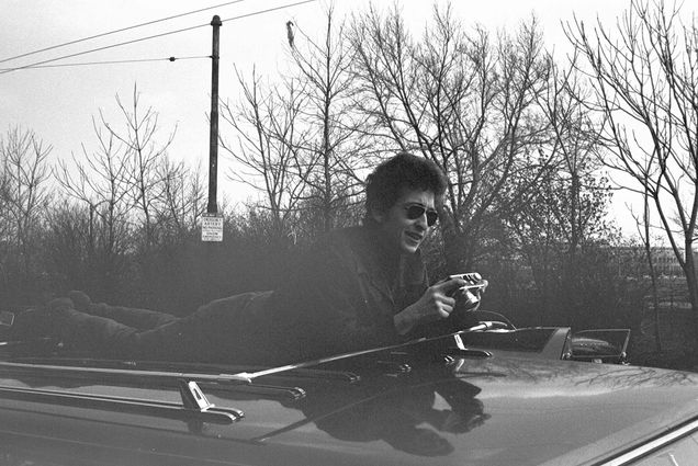 a black and white photo of Dylan clowning atop a car parked on Memorial Drive in April 1964. He lies on top of care and looks down at a camera in his hands.