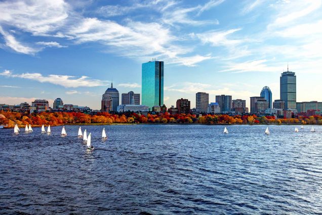 Photo taken on the Charles River, looking from the Longfellow Bridge down river towards the Prudential. A group of white sail boats are seen, and the trees along the river have autumn-colored leaves.