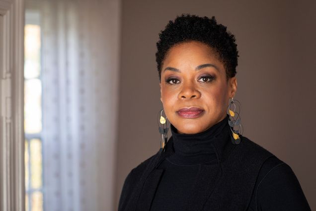 photo of Crystal Williams posing in her home. She looks at the camera and wears a black turtleneck and intricate, dangling blue and gold earrings