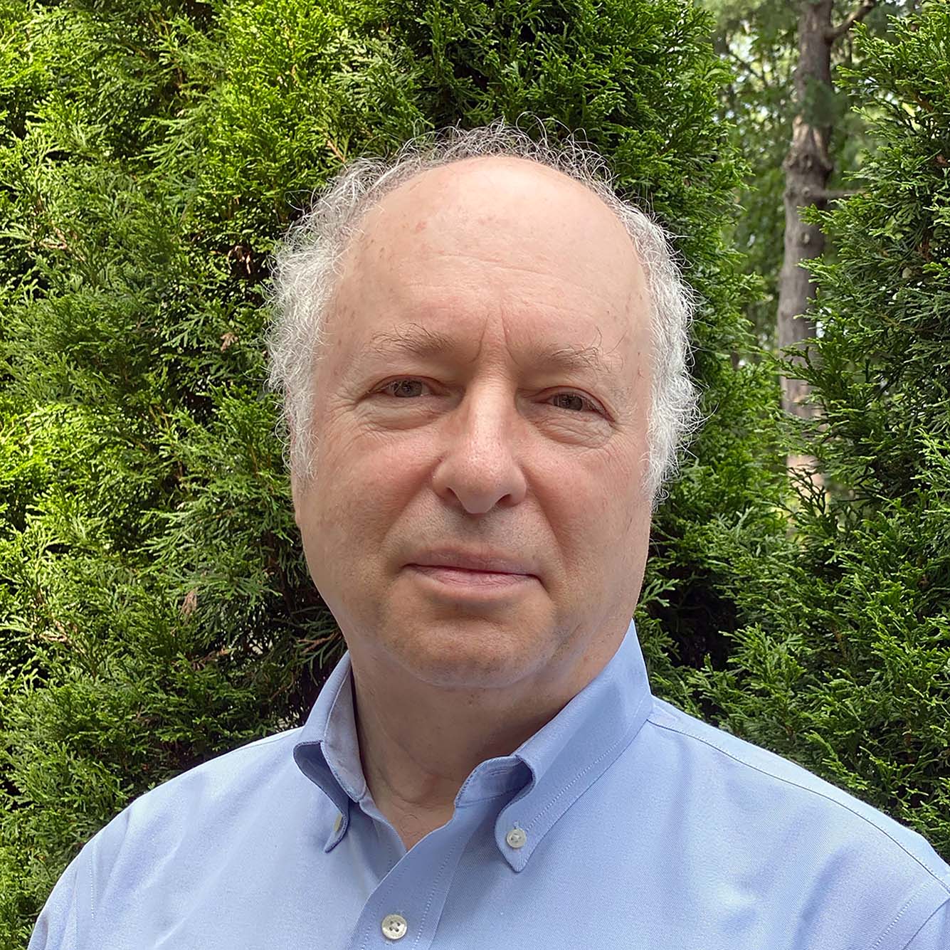 headshot of Greg Blonder, visiting scholar and former professor of the practice in mechanical engineering at BU’s College of Engineering. He smiles as he looks to the camera wearing a light blue collared shirt. Background is full of trees and greenery.