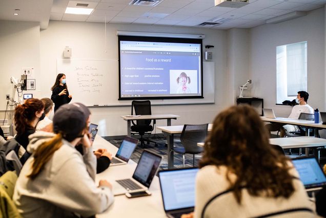 Photo of Bianca Saffiro (CAS’24), at the front of the class in black, presenting her final project, “Eating and Psyche: Food as Emotional Compensation,” to the class on December 1. In the foreground, students with their laptops open are seen.