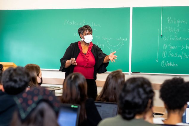Photo of Margarita Guillory, wearing a white face mask, red shirt and black jacket, lecturing at the front of her Religion and Hip Hop course. She extends her hand toward the class and the backs of students' heads are seen.