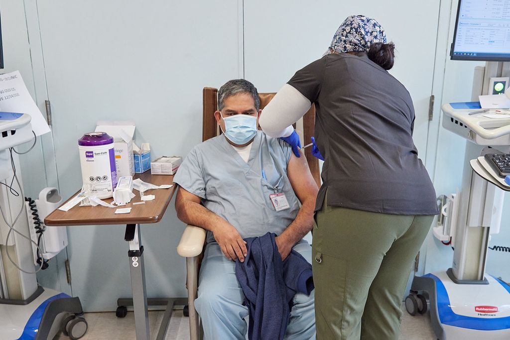 Photo of a healthcare worker giving a coronavirus vaccine to a fellow healthcare worker who sits in a chair and wears scrubs and a face mask.
