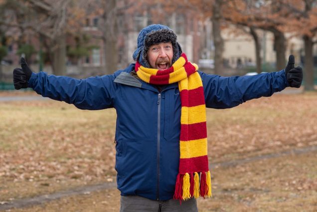 Photo of Hutch Hutchinson, a veteran New England outdoorsman, wearing a yellow and red scarf, blue hat with panels, and blue winter coat. He smiles, extends his arms joyfully, as he explains tips for how to dress for winter.