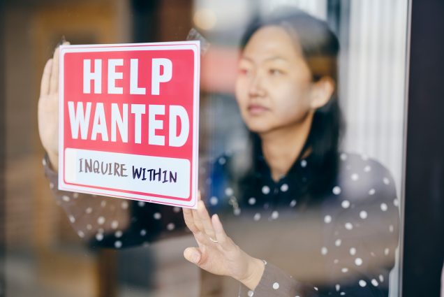 Photo of an Asian woman business owner putting up a help wanted sign in her store window