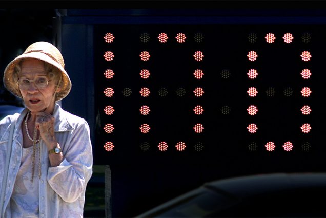A woman in a hat stands in front of an electronic billboard that reads 100 degrees