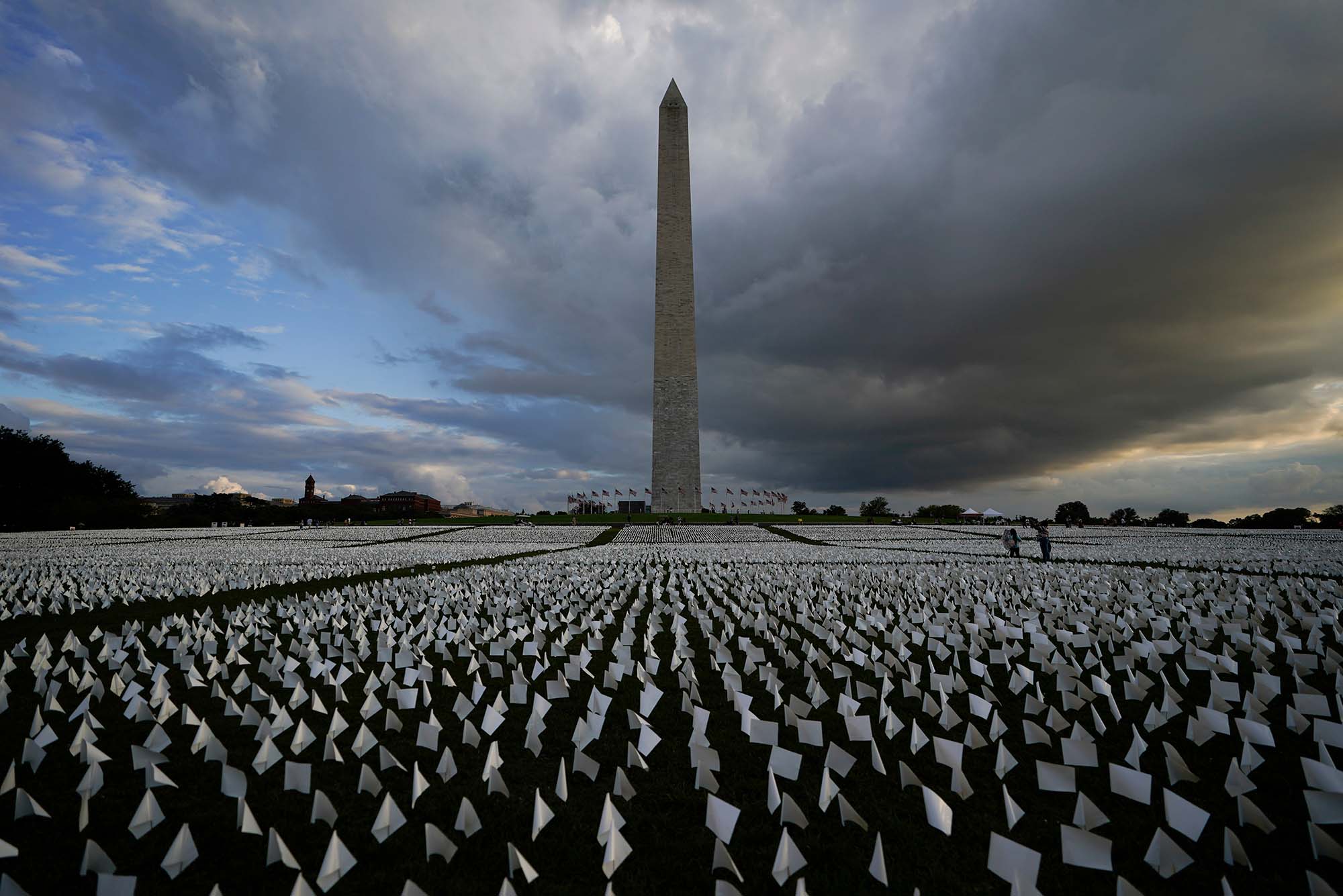 Photo of a field of white flags around the Washington Monument. Artist Suzanne Brennan Firstenberg’s temporary art installation on the National Mall, In America: Remember, features white flags that symbolize a life lost to COVID-19. Behind the mall, the sky is ominous and dark.