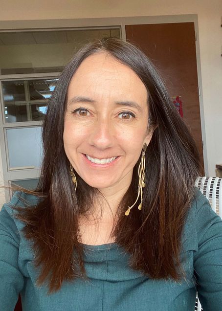 Photo of Patricia Cortes, a woman with shoulder length brown hair who smiles, and wears long dangly errands and a mauve green sweater.